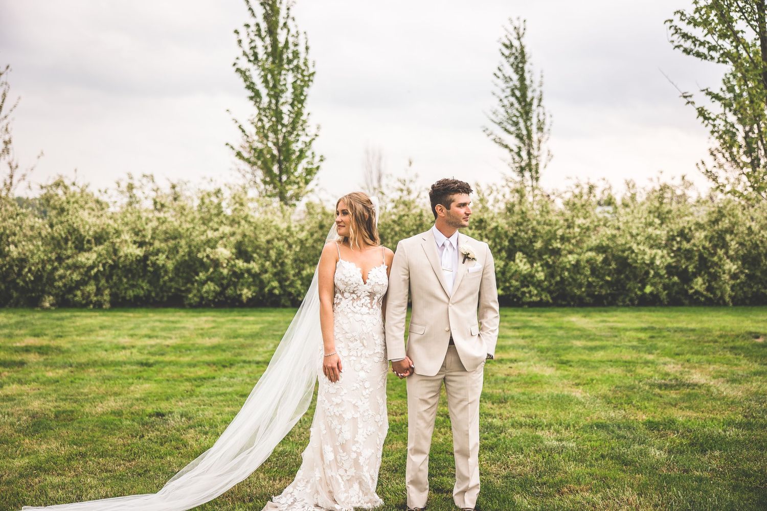 Wedding couple posing outdoors in a green field with flowing veil and light-colored attire.