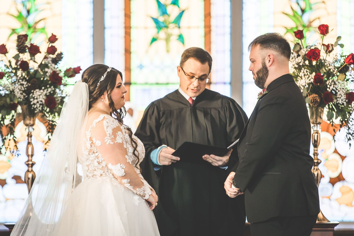 Wedding ceremony taking place at altar decorated with red roses and greenery.