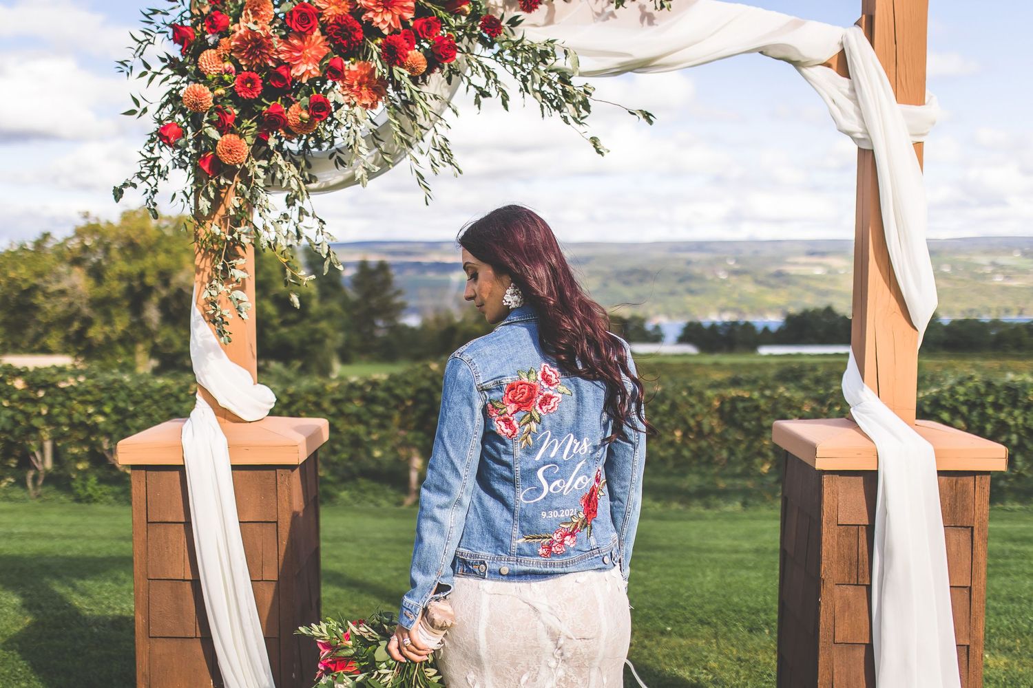 Wedding couple poses in outdoor garden setting with red floral arch and mountain views in background.