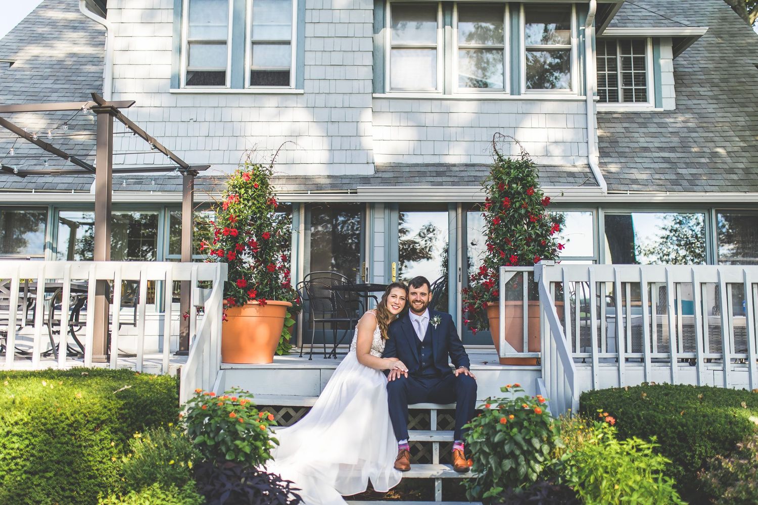 Wedding portrait on front steps of gray shingled house with potted flowers.
