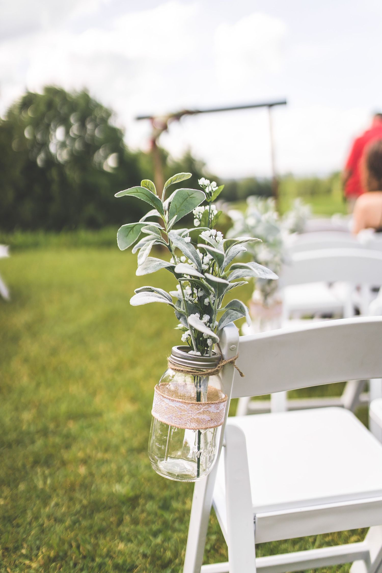 Mason jars with small green plants line white chairs along a grassy outdoor aisle.
