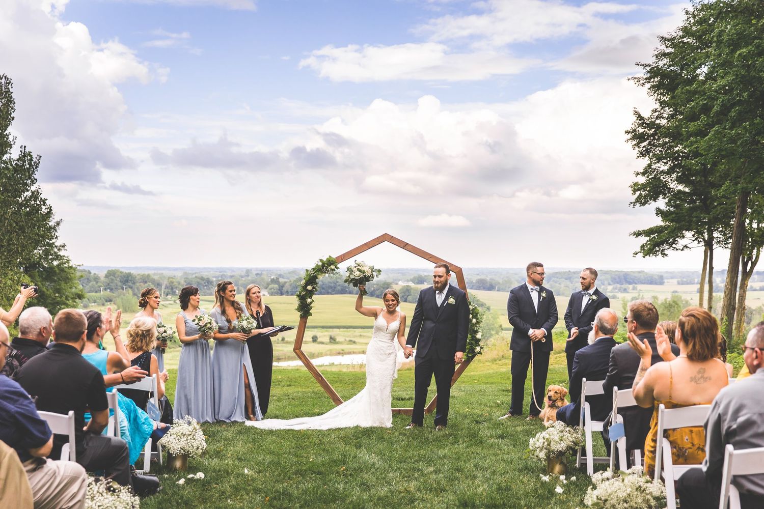 Outdoor wedding ceremony setup with wooden hexagonal arch and guests seated on white chairs.