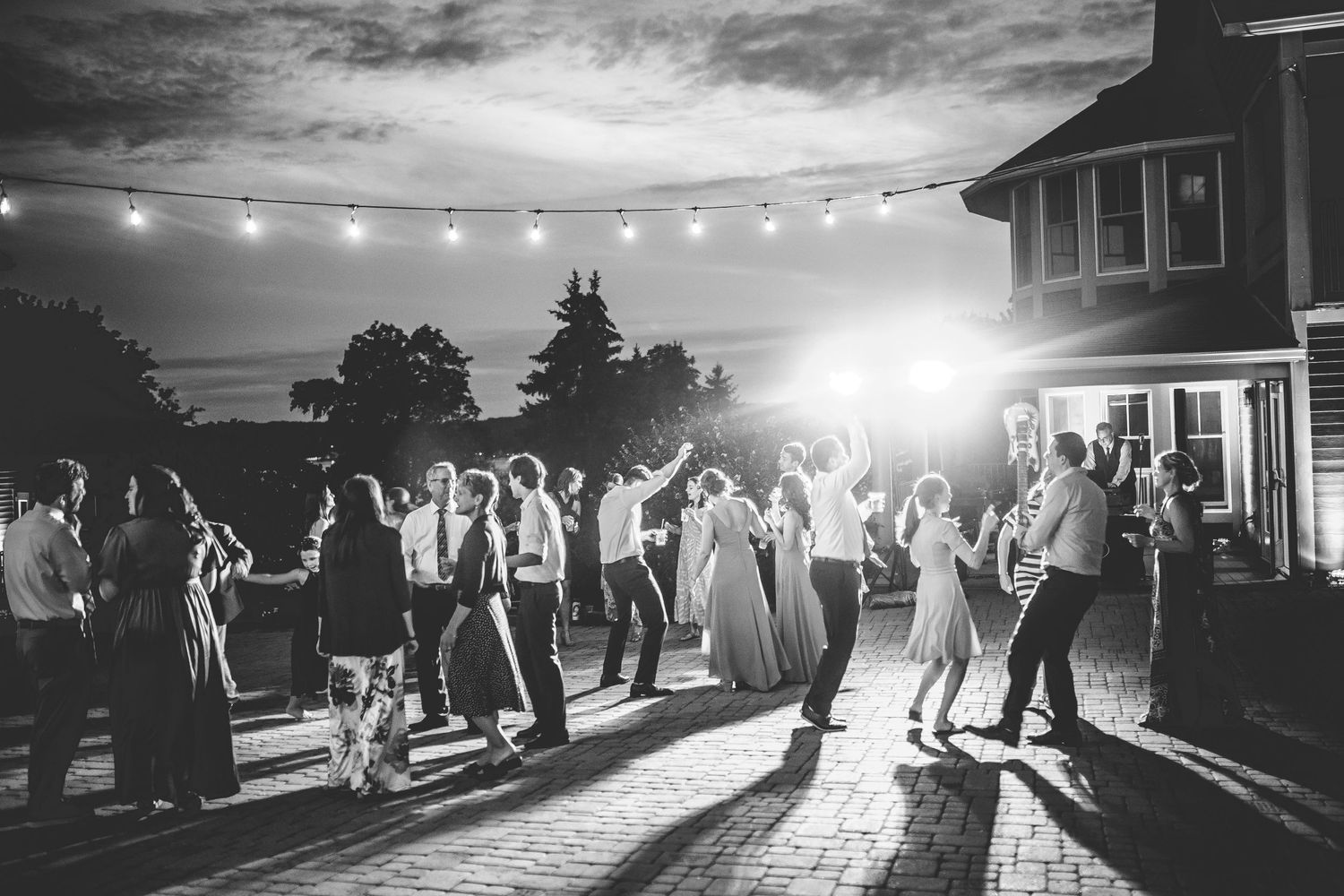 String lights illuminate an outdoor reception area as guests dance at dusk.