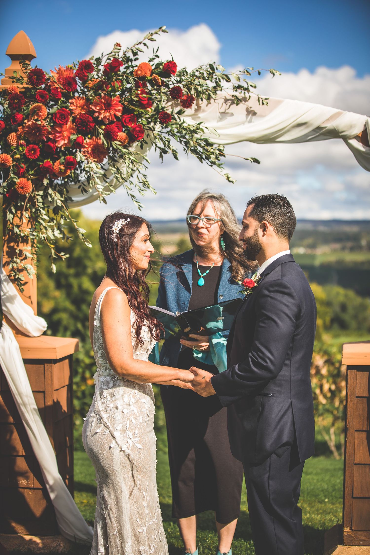 Outdoor wedding ceremony under blue skies with red floral arch decorations.