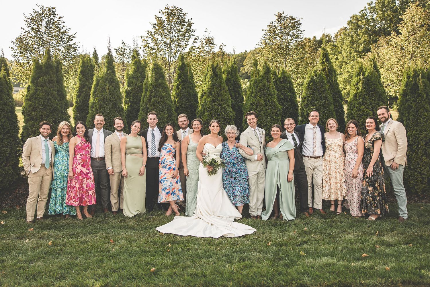 Large wedding party poses for group photo outdoors with evergreen trees in background.