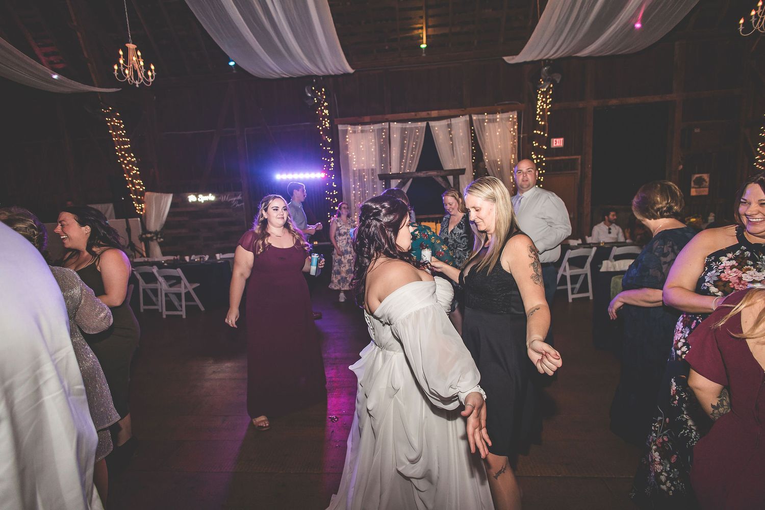 Wedding reception guests dance under mood lighting and white fabric draping in a rustic venue.