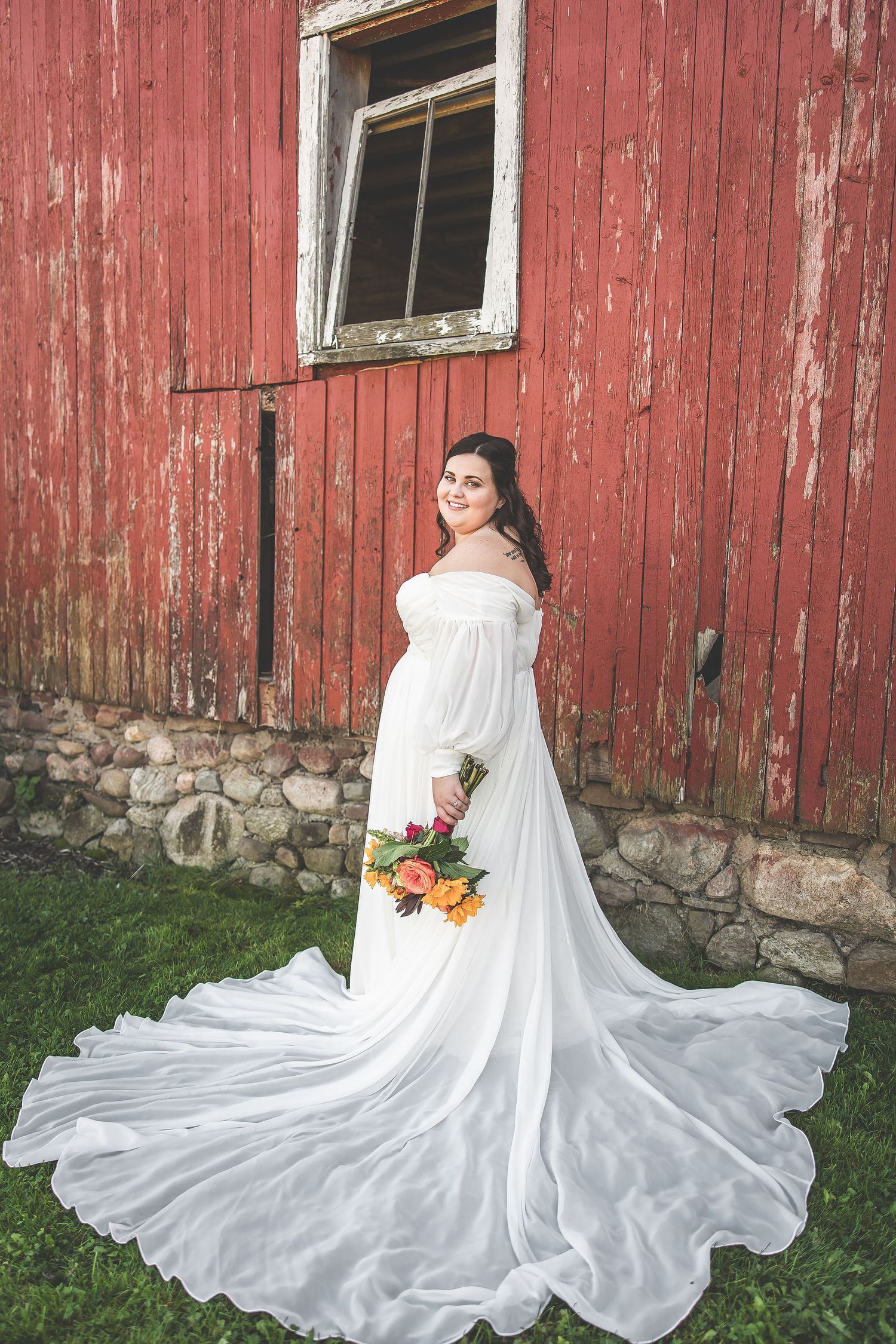 Bride in off-shoulder wedding gown with flowing train poses by rustic red barn.