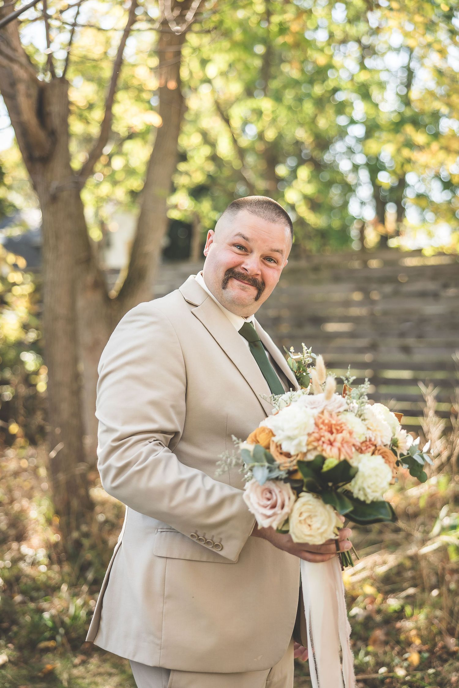 Groomsman in tan suit holding a peach and white floral bouquet in an outdoor garden setting.