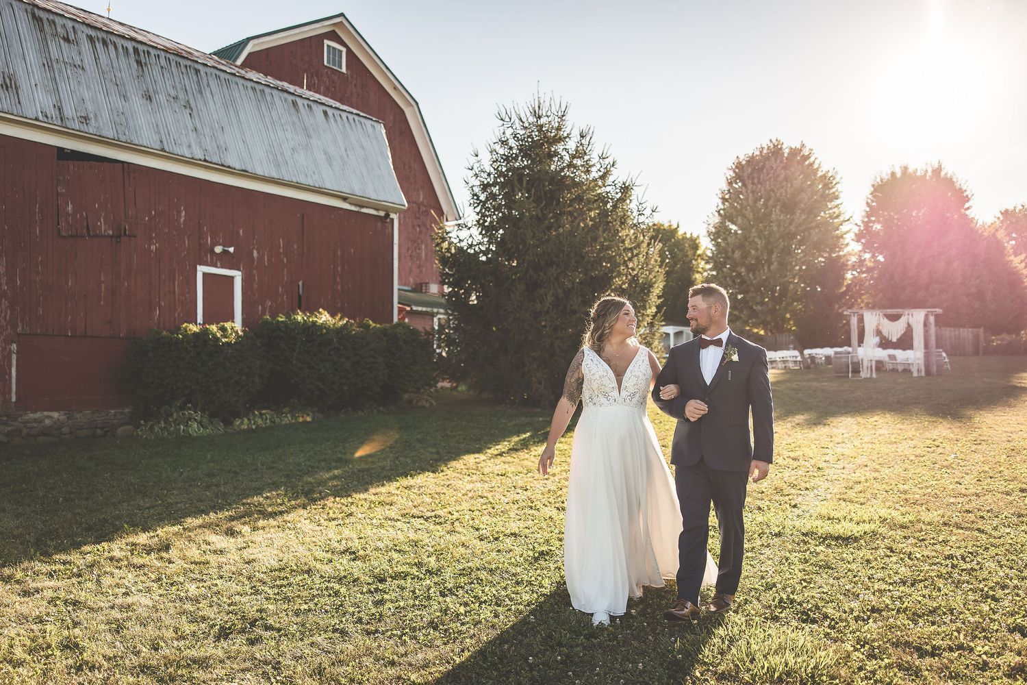 Wedding couple walks near red barn during sunset with dramatic shadows.