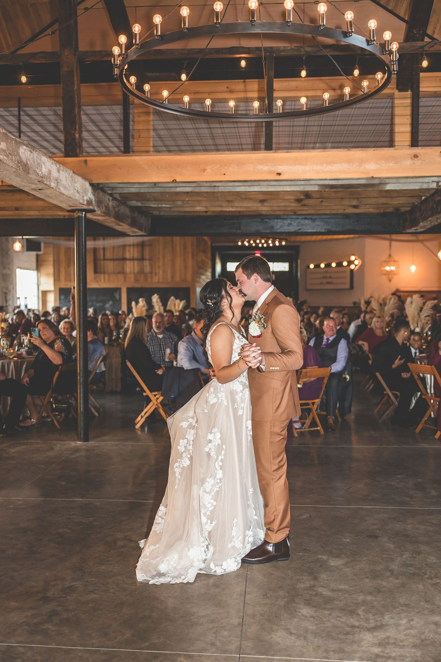 A couple performs their wedding dance in a rustic venue with wooden beams and string lights.