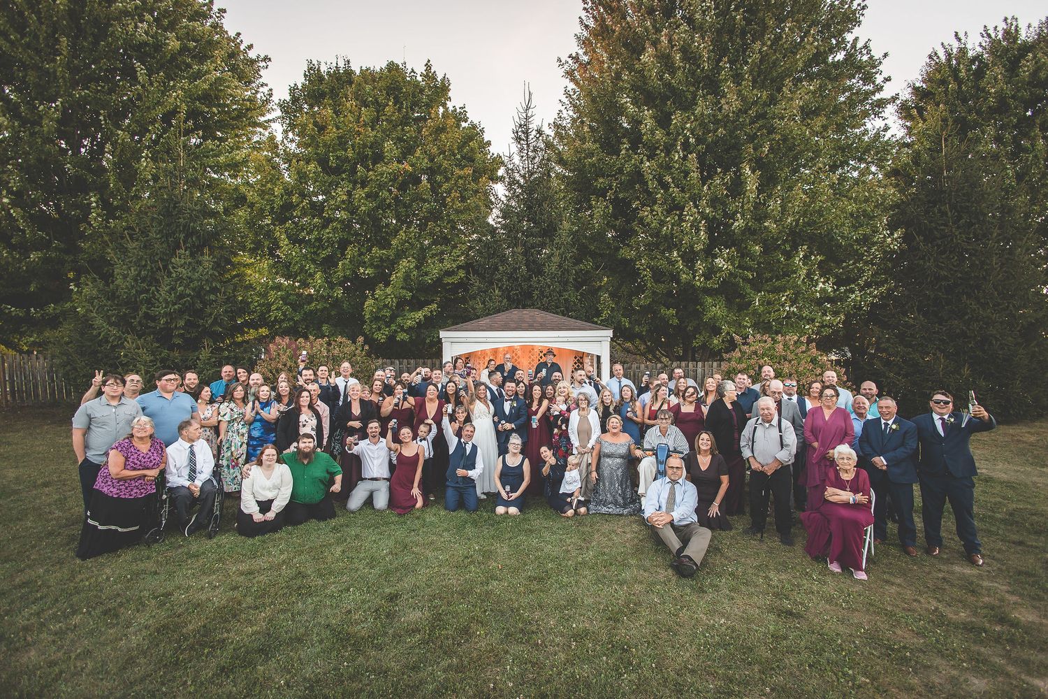 Large group wedding photo of guests and wedding party on lawn in front of trees.