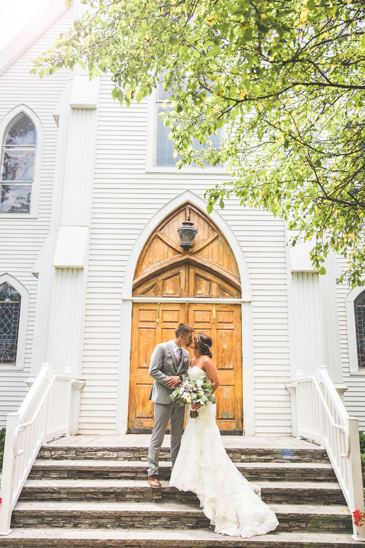 Couple poses on church steps in front of wooden arched door with white exterior and stained glass windows.