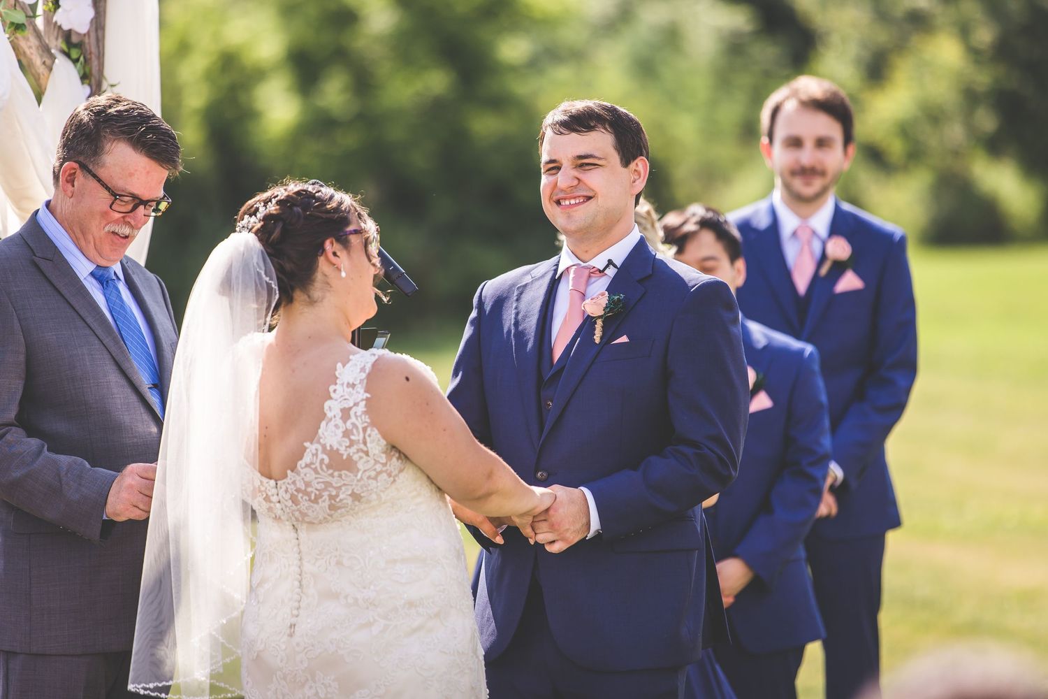 Wedding ceremony participants in navy suits and formal attire gathered outdoors on a sunny day.