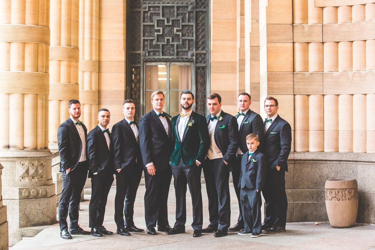 Groomsmen in formal suits lined up against stone pillars.
