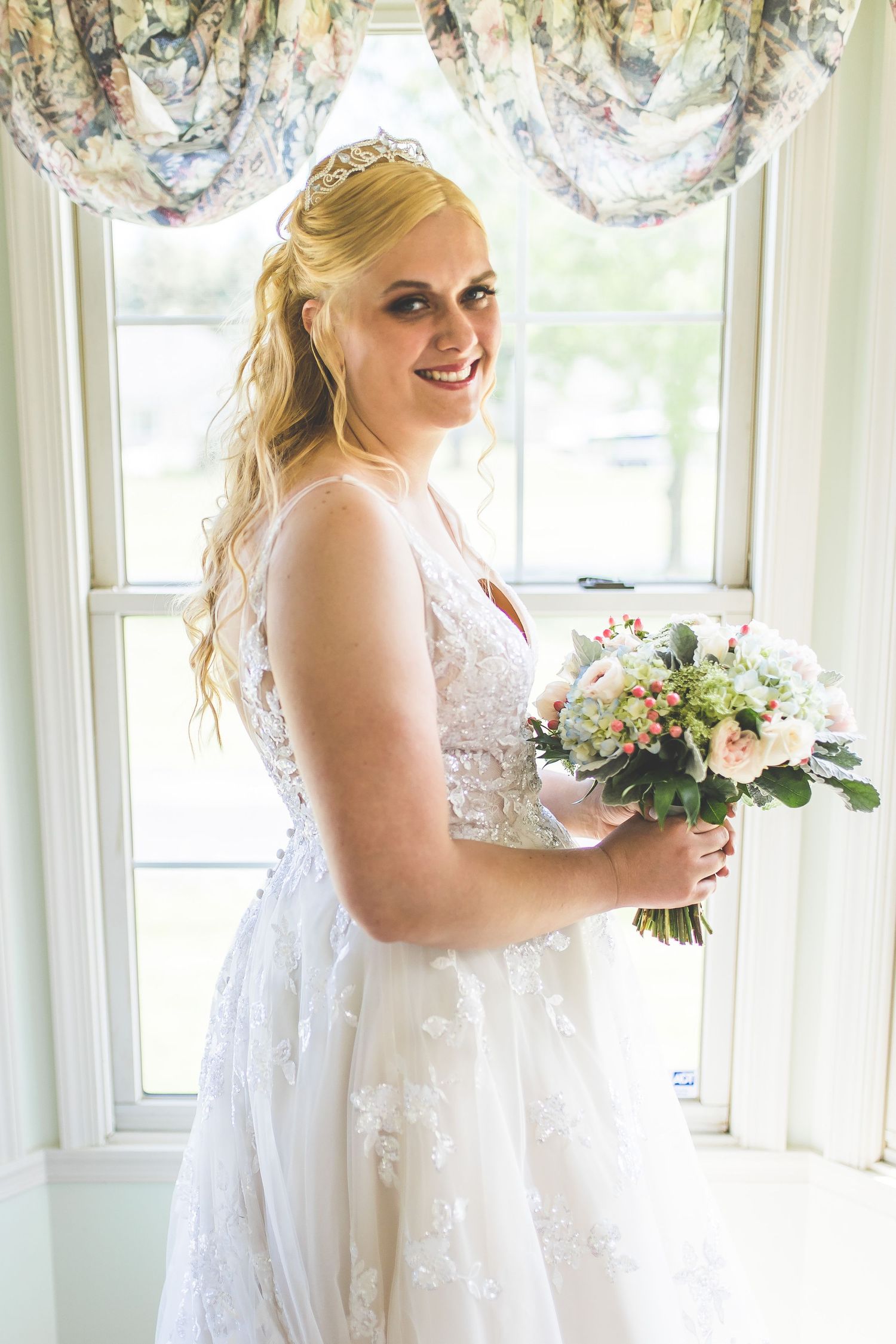 A bride in a white lace dress holds a bouquet in a bright sunlit room.