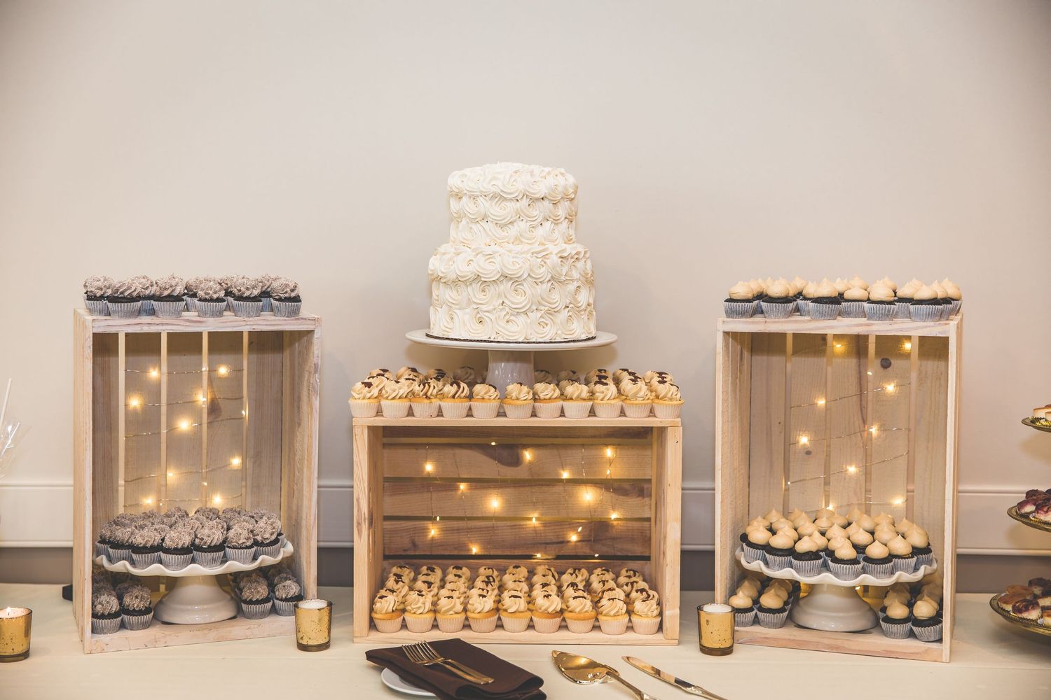 Elegant dessert table display with wedding cake and cupcakes illuminated by string lights.