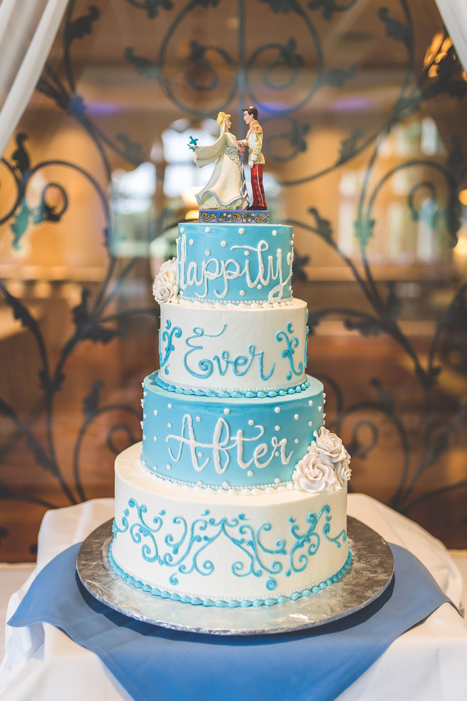 Three-tier wedding cake decorated in blue and white with 'Happily Ever After' theme.