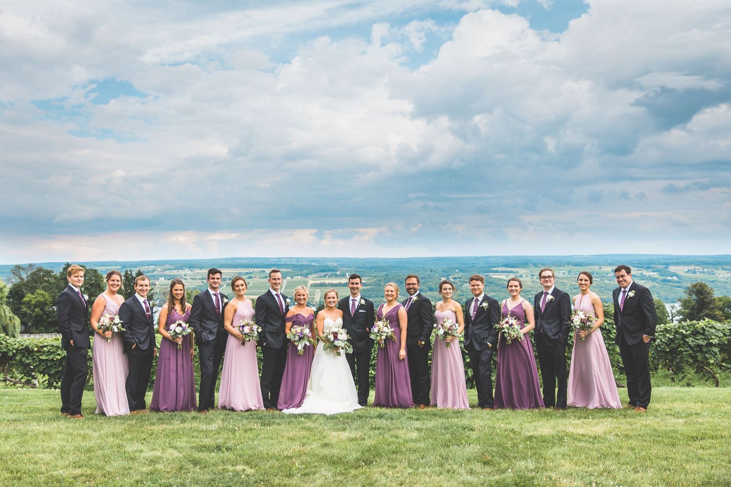 Wedding party in purple and pink attire posing together on a hilltop with scenic backdrop.