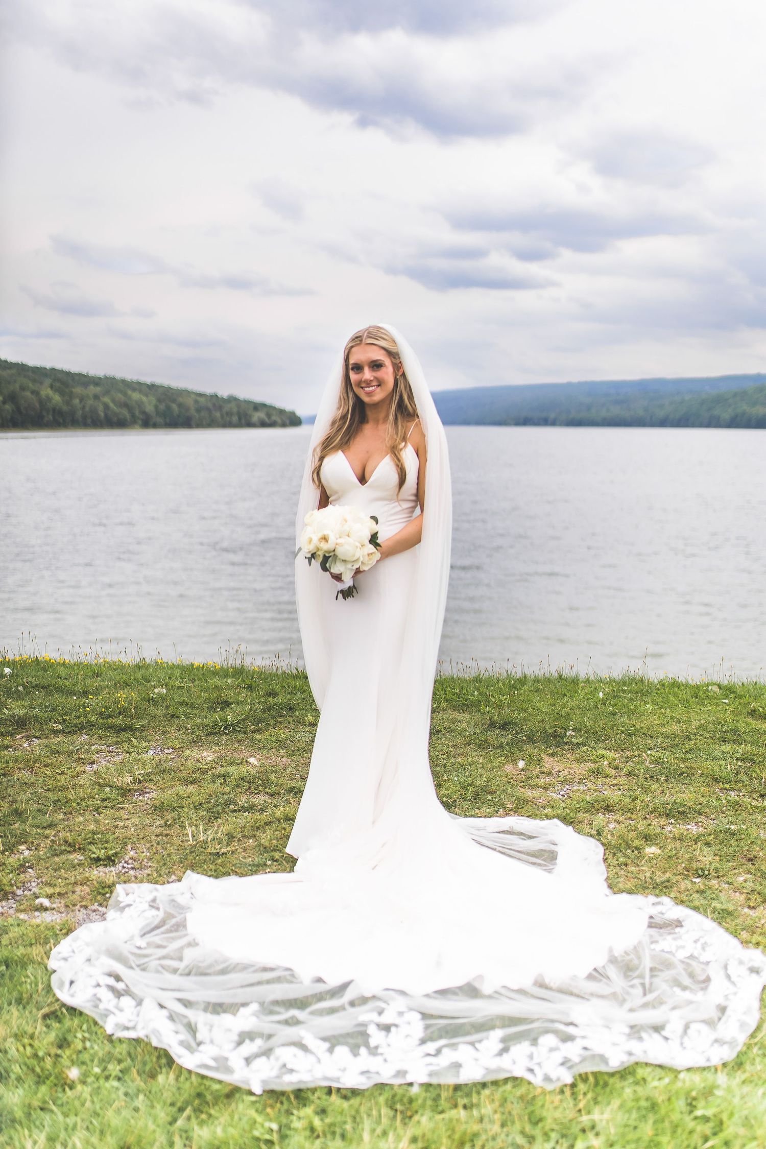 A flowing white wedding dress with long train spreads across green grass near a serene lake with mountains in background.