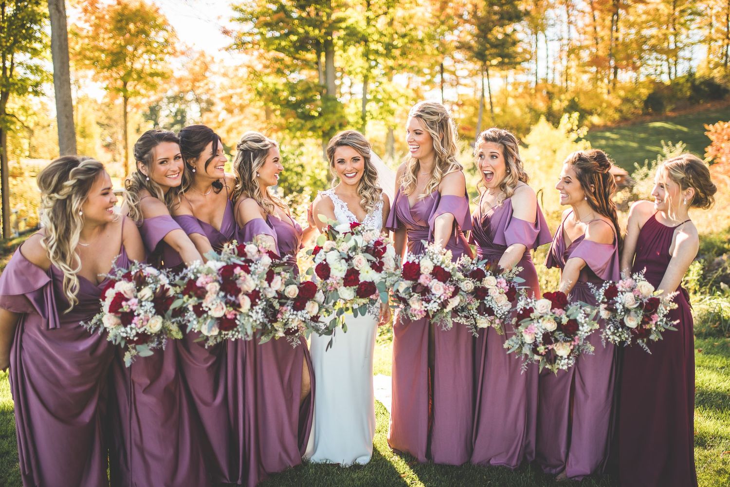 Wedding party in purple dresses poses outdoors during golden hour with bouquets.