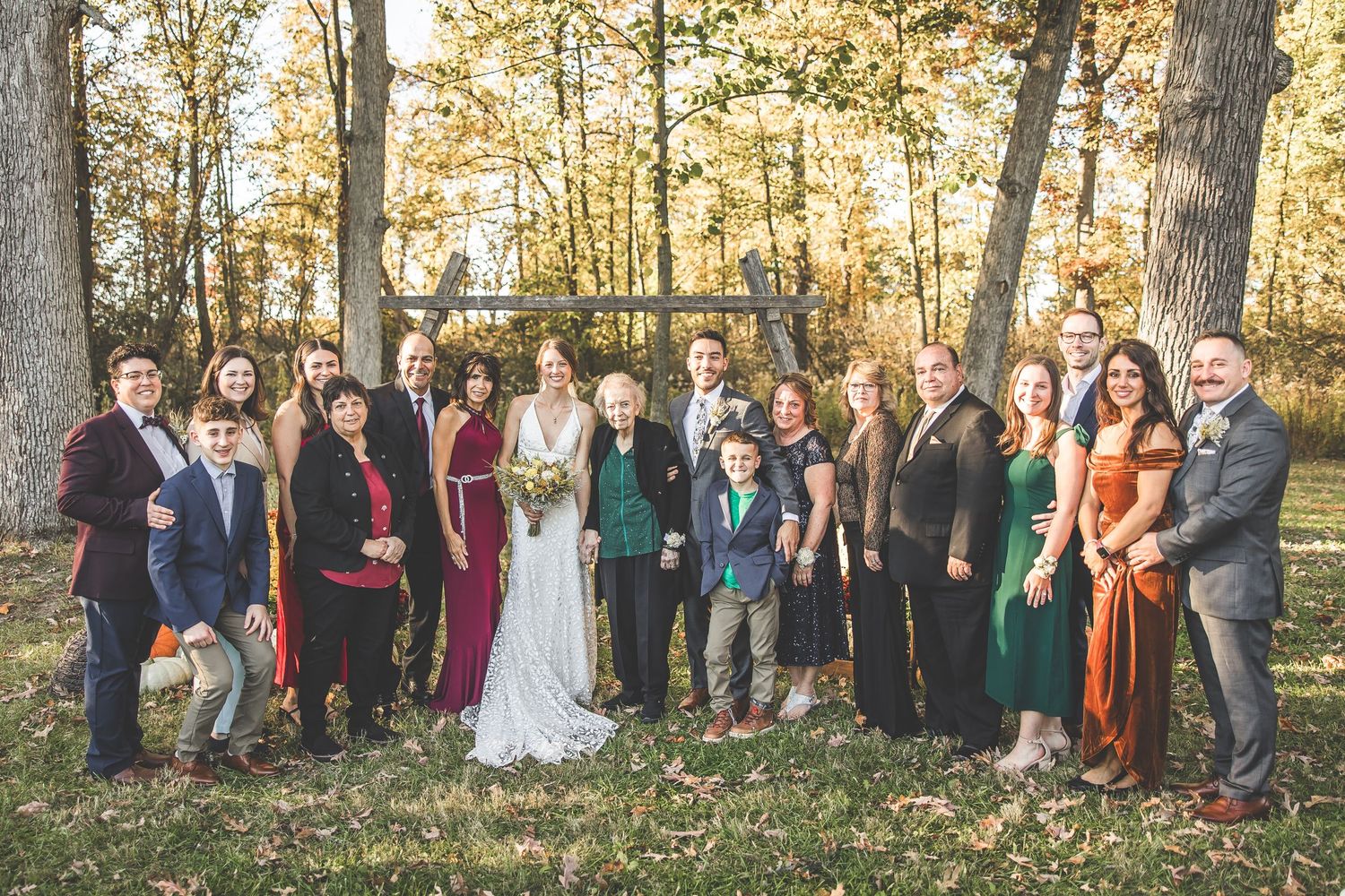 Large wedding party group photo with formal attire against an autumn forest backdrop.