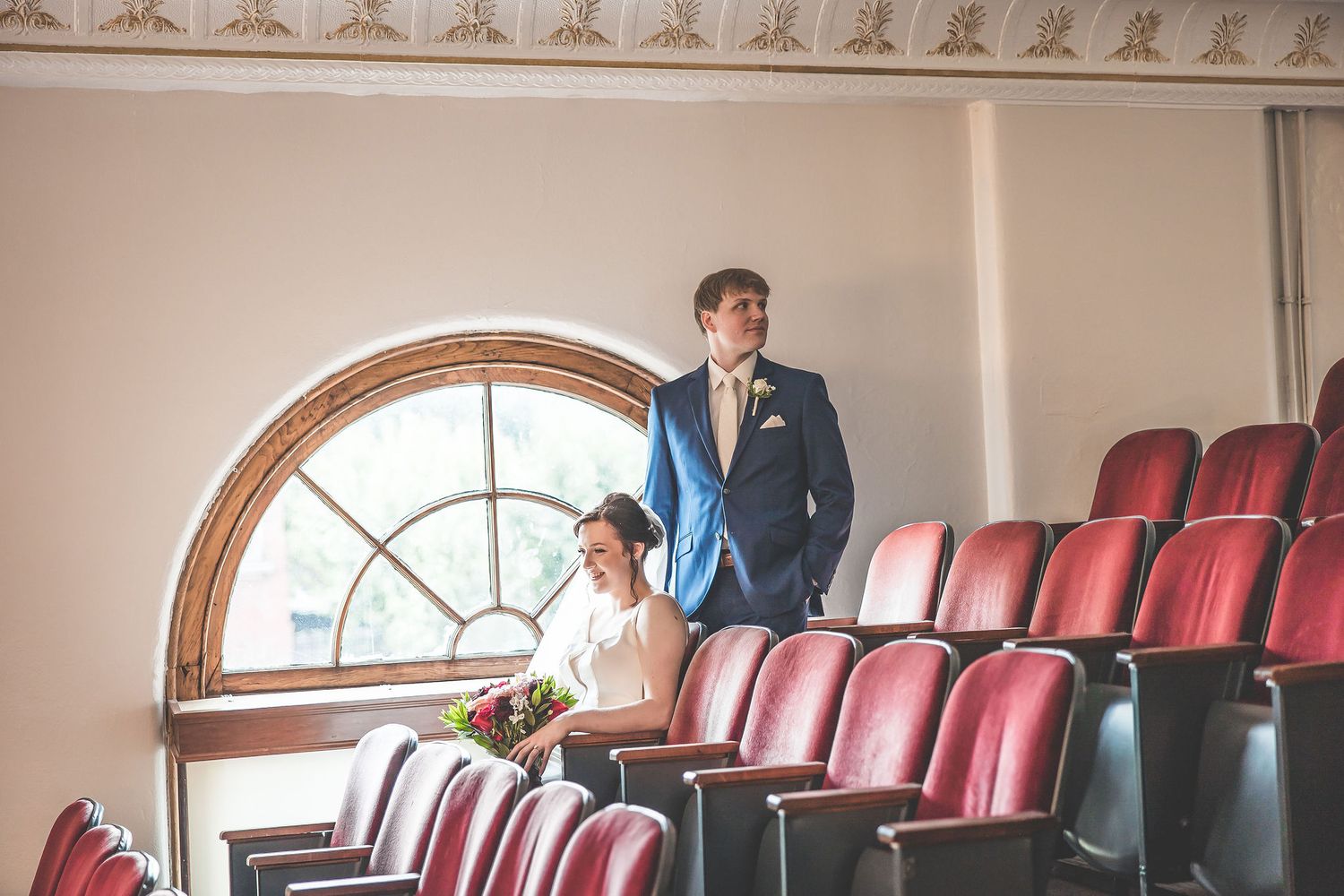 Historic theater balcony with decorative molding and red velvet seats sets the stage for wedding photos.