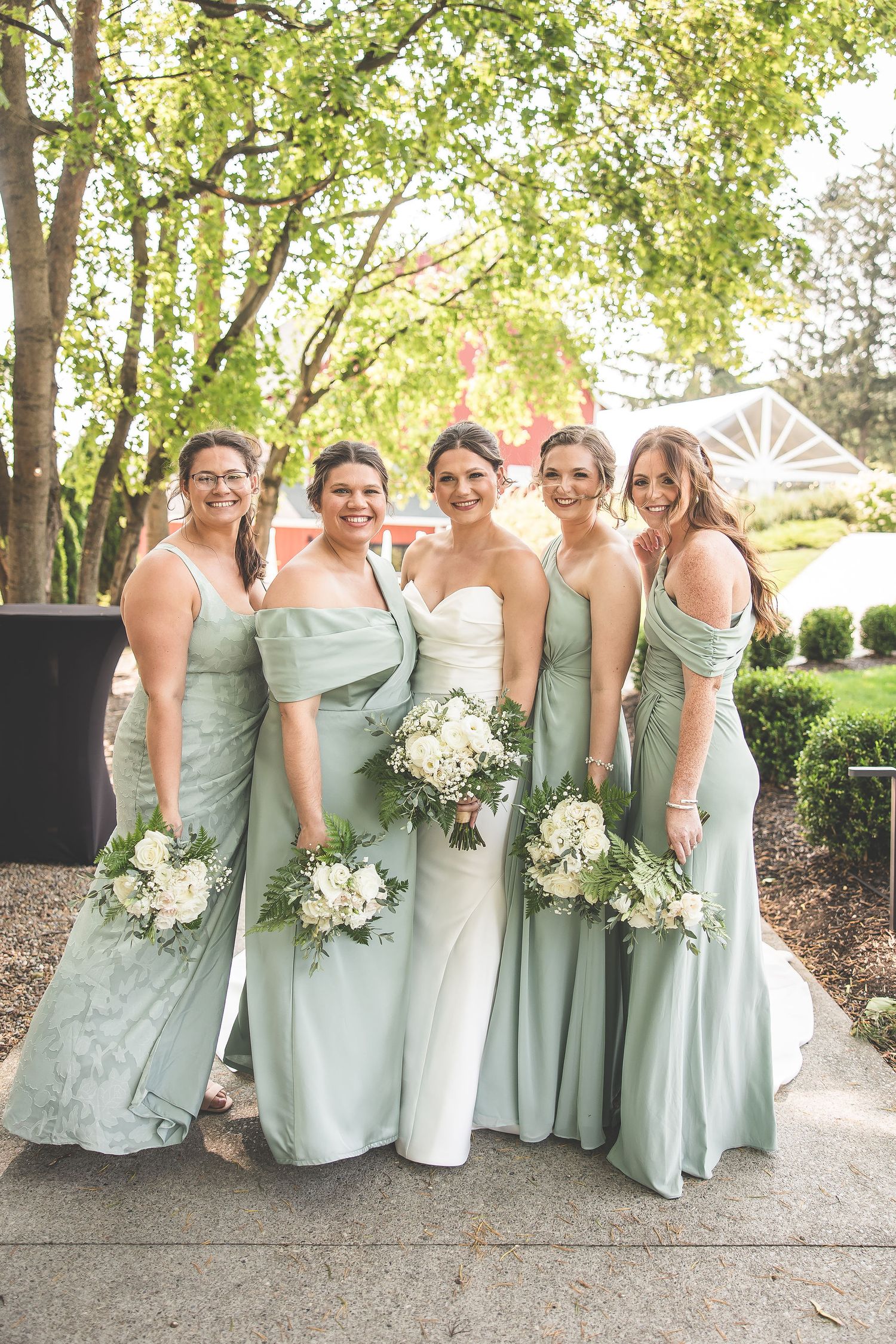 Bridesmaids in soft sage green dresses pose together while holding white floral bouquets in an outdoor garden setting.
