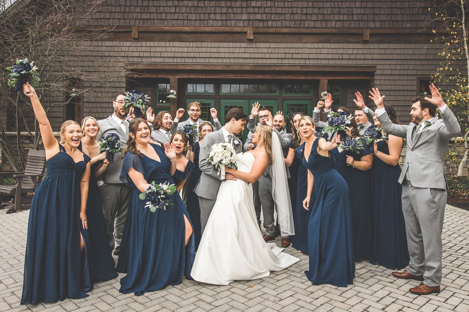 Wedding party members cheer and pose energetically outside brick building.