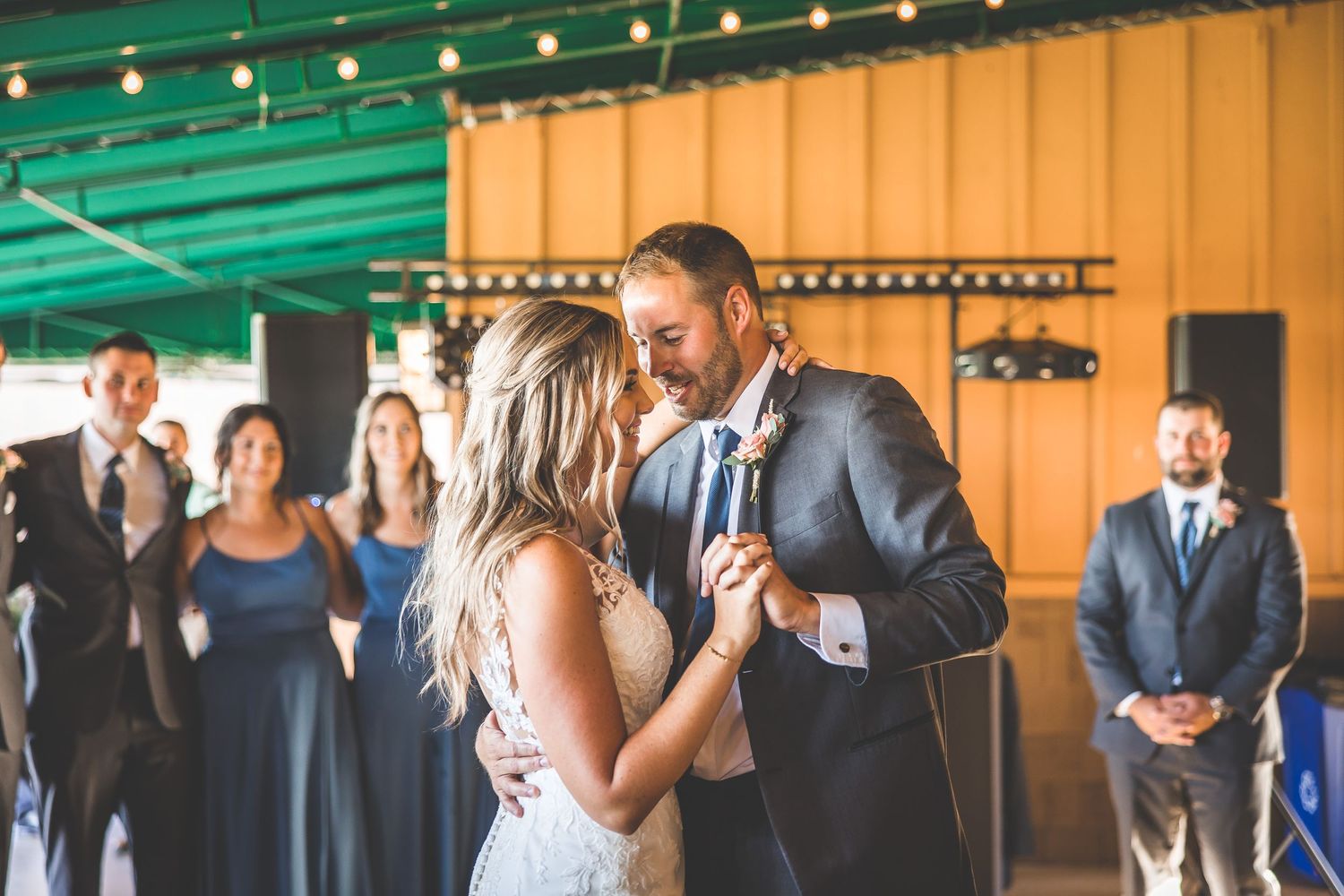 Wedding guests watch a couple dance under string lights in a rustic venue with orange walls.