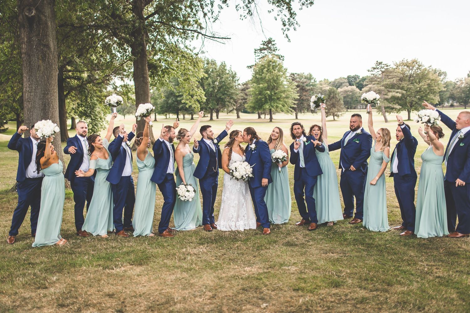 Wedding party in blue dresses and suits poses playfully on a grassy hill.