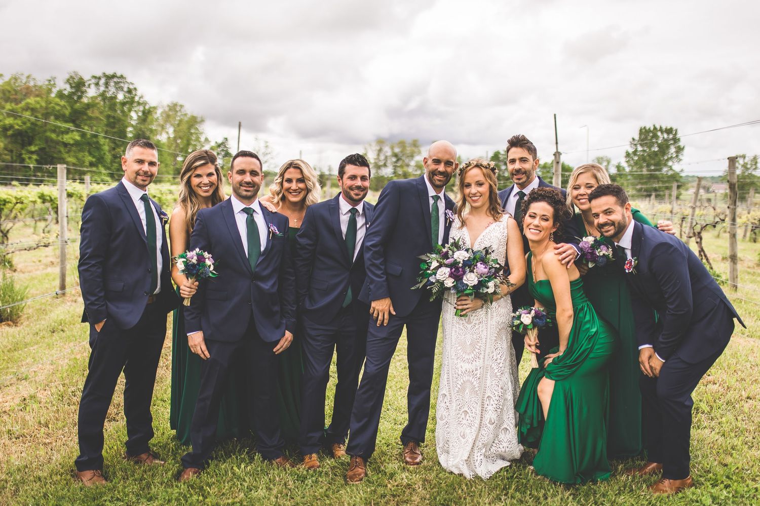 Wedding party in navy suits and emerald green dresses poses together outdoors in a rustic vineyard setting.