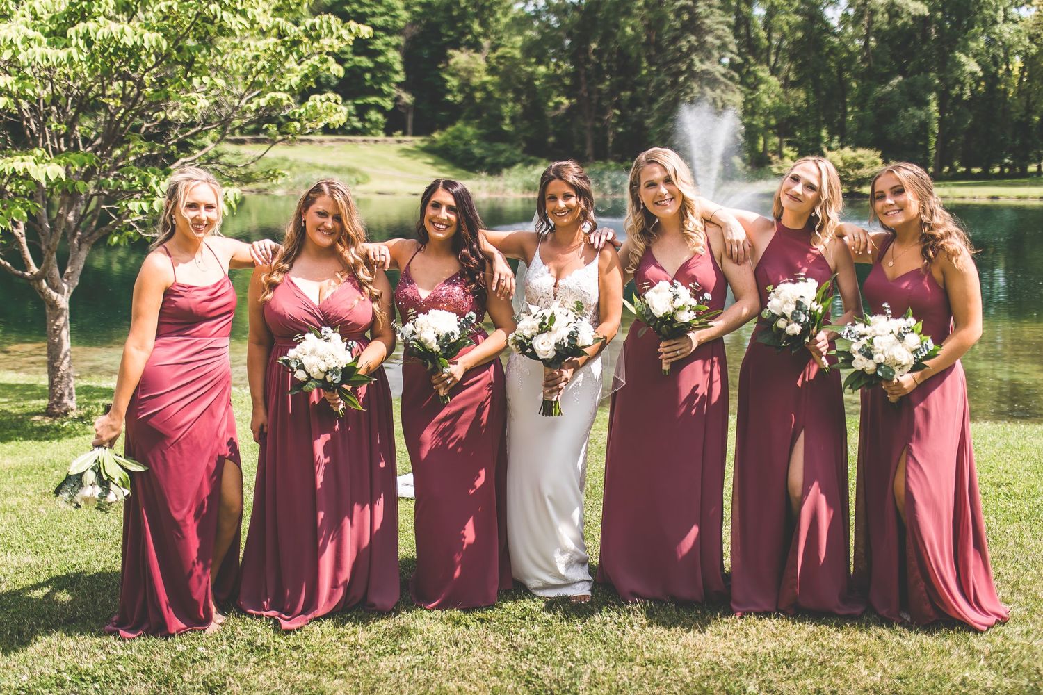 Wedding party members in burgundy dresses pose together with bouquets by a lake.