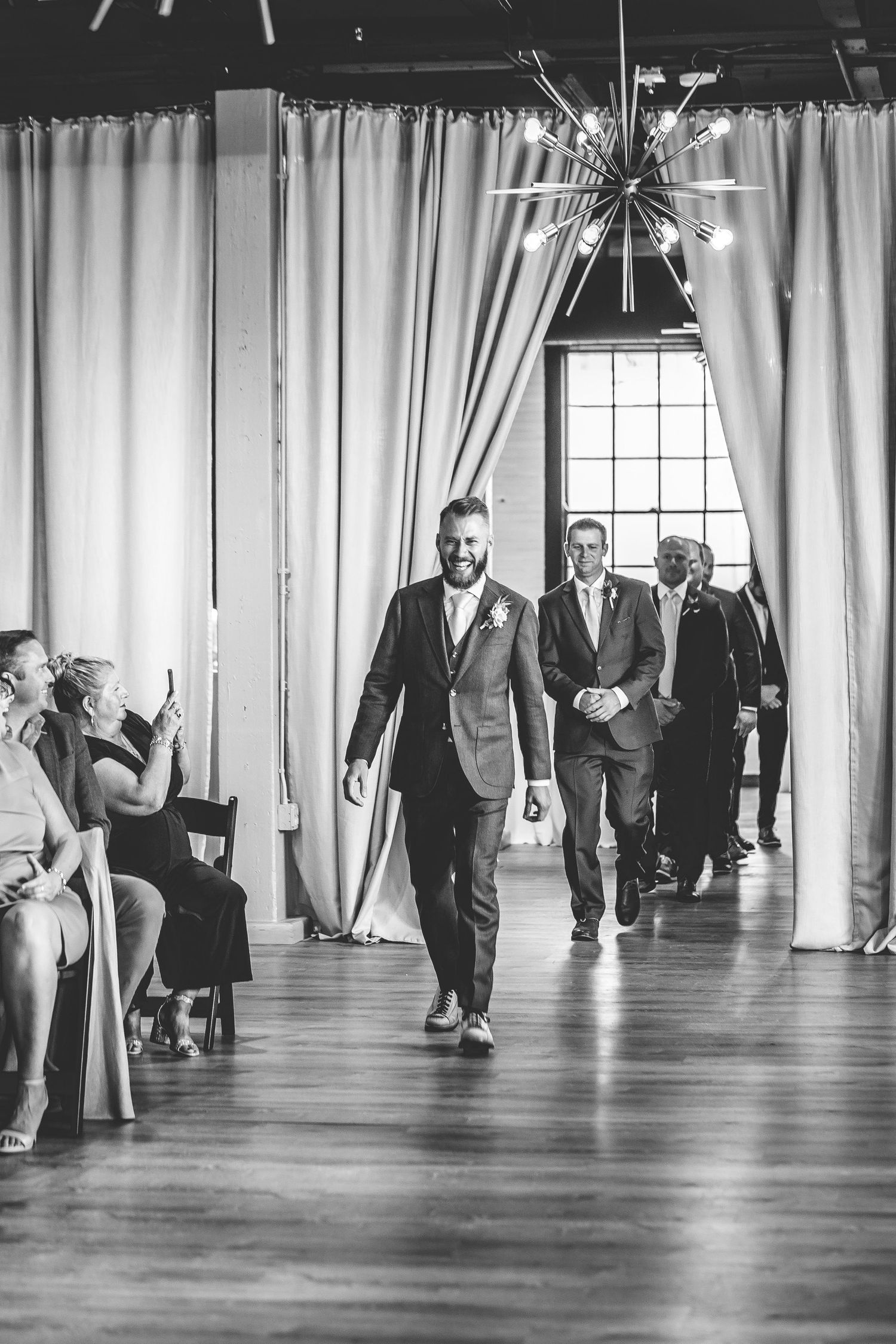 Black and white photograph of wedding party members walking through an elegant venue hallway.