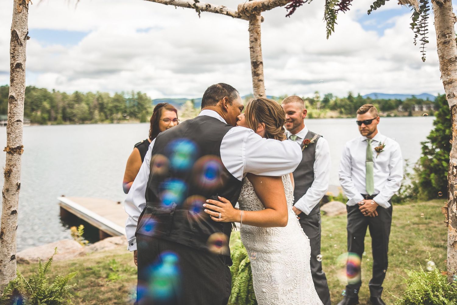 Outdoor lakeside wedding ceremony under a rustic birch arch with guests looking on during an intimate moment.