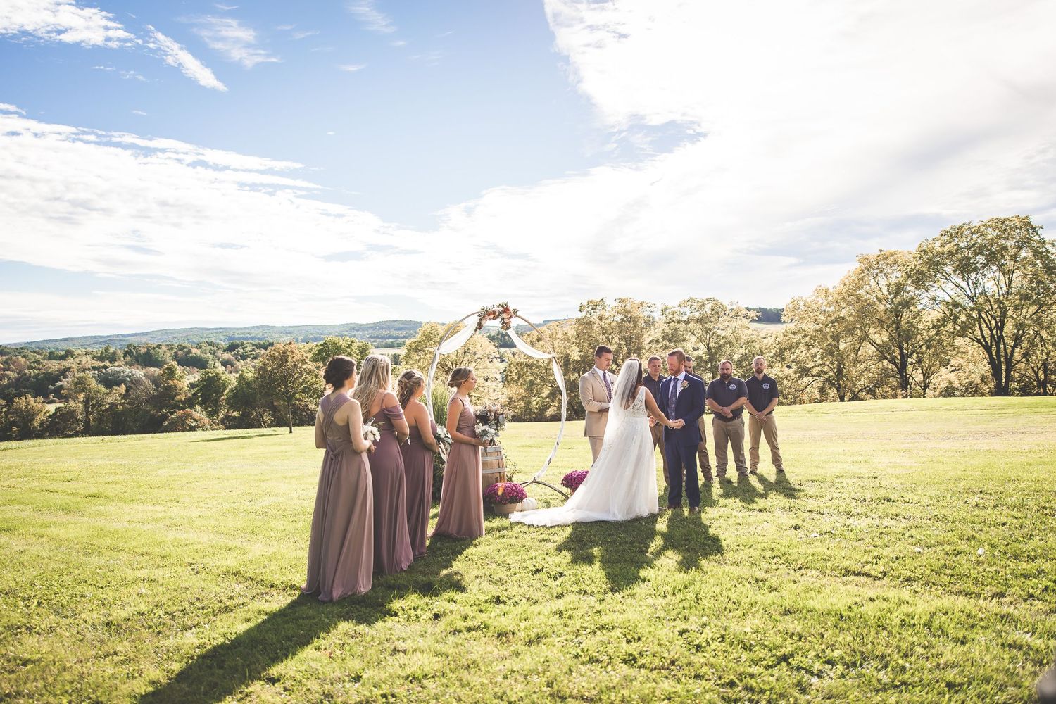 Wedding party and guests gathered on a hilltop during a sunny outdoor ceremony with scenic views.
