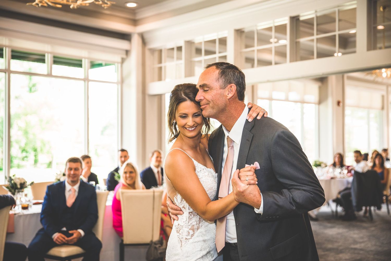 Couple shares romantic dance moves in a bright reception hall with guests looking on.