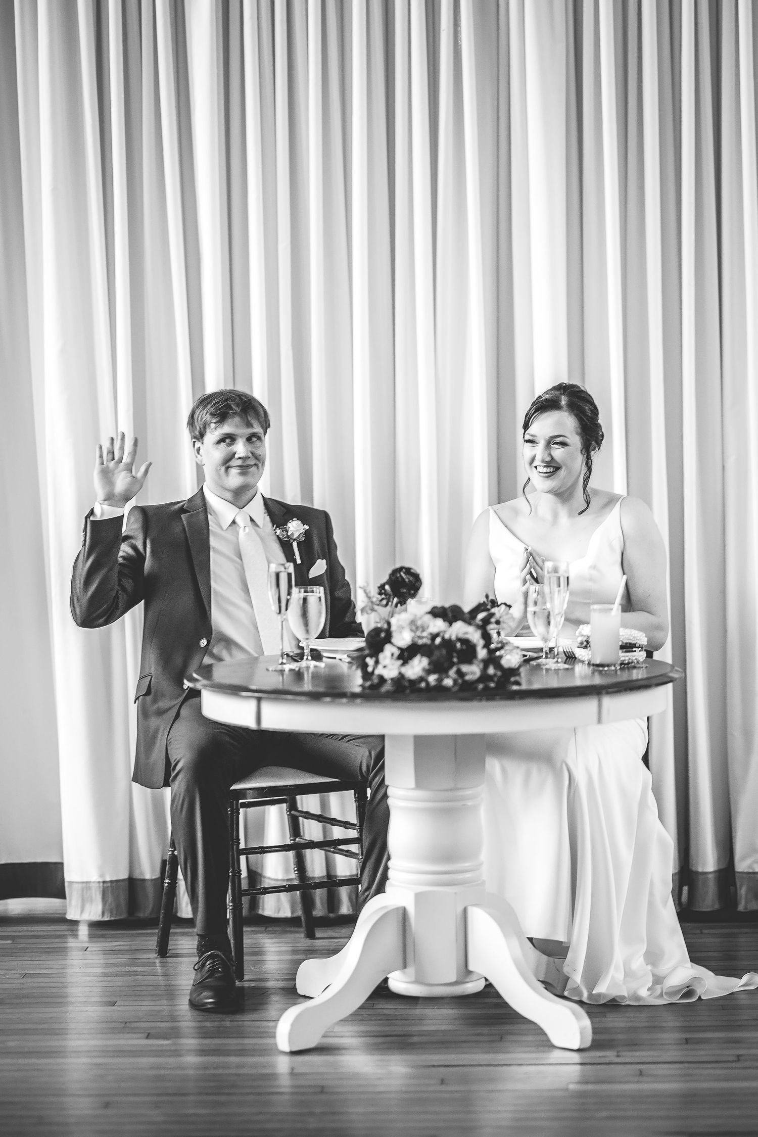 A black and white reception photo showing a couple seated at their sweetheart table.