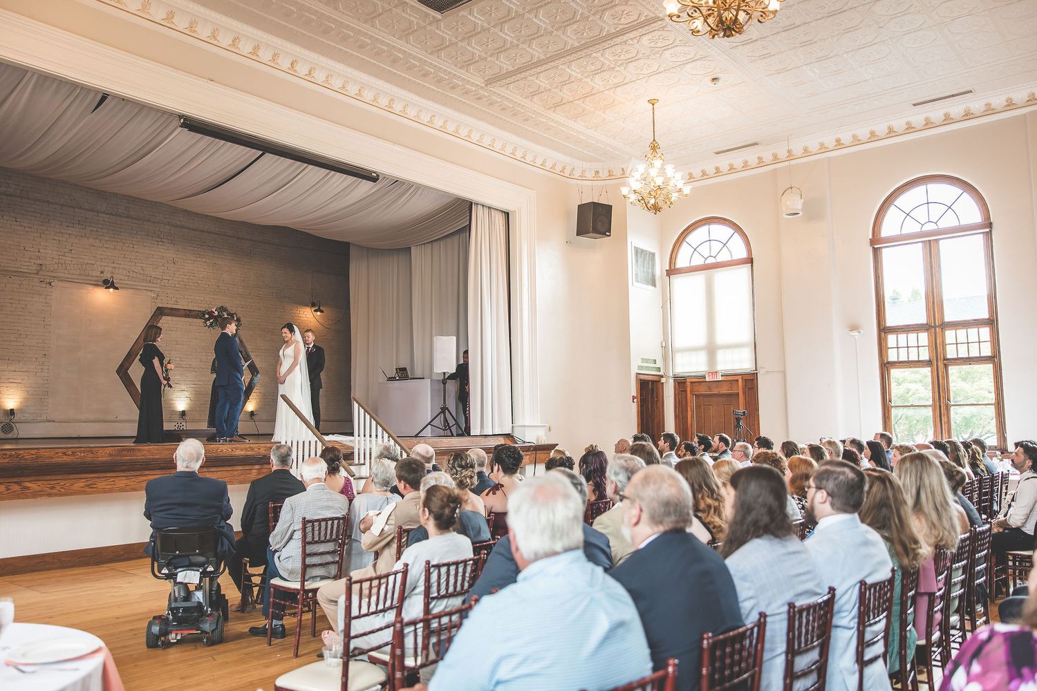 Guests seated in white chairs during indoor wedding ceremony in historic venue with ornate ceiling.