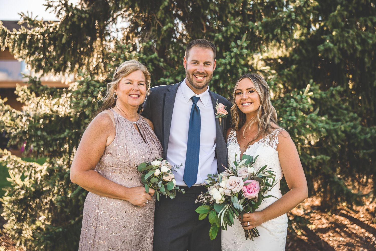 Outdoor wedding portrait with couple and guest in formal attire against sunlit trees.