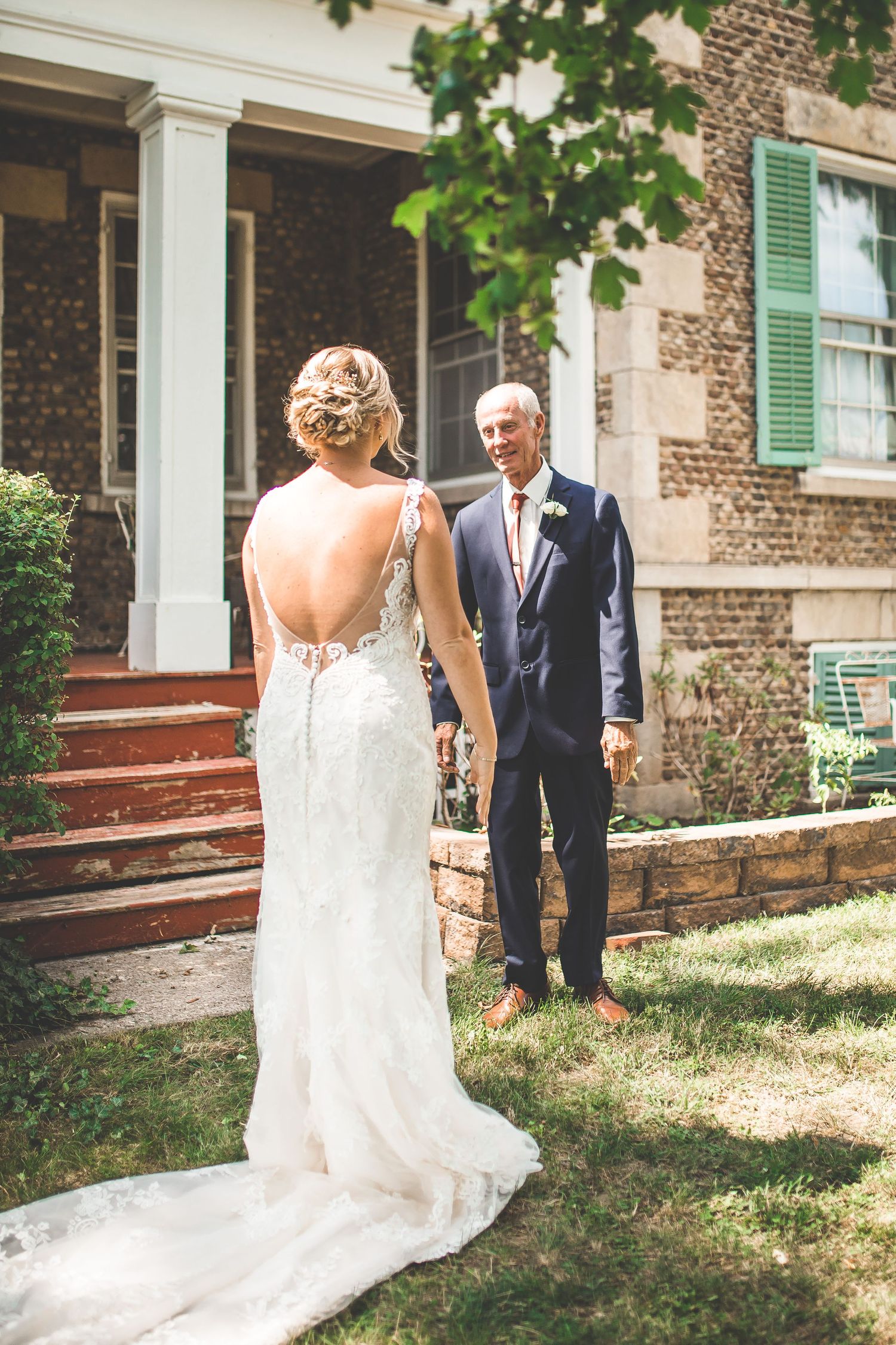 A bride in a backless gown stands on outdoor steps during a first look moment.