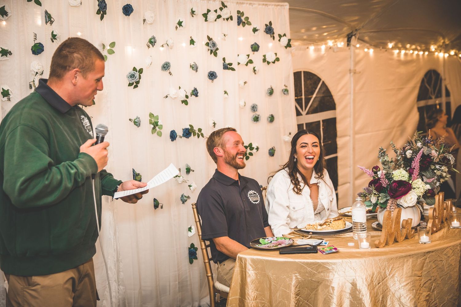 Wedding reception space with white fabric backdrop adorned with cascading greenery and string lights.