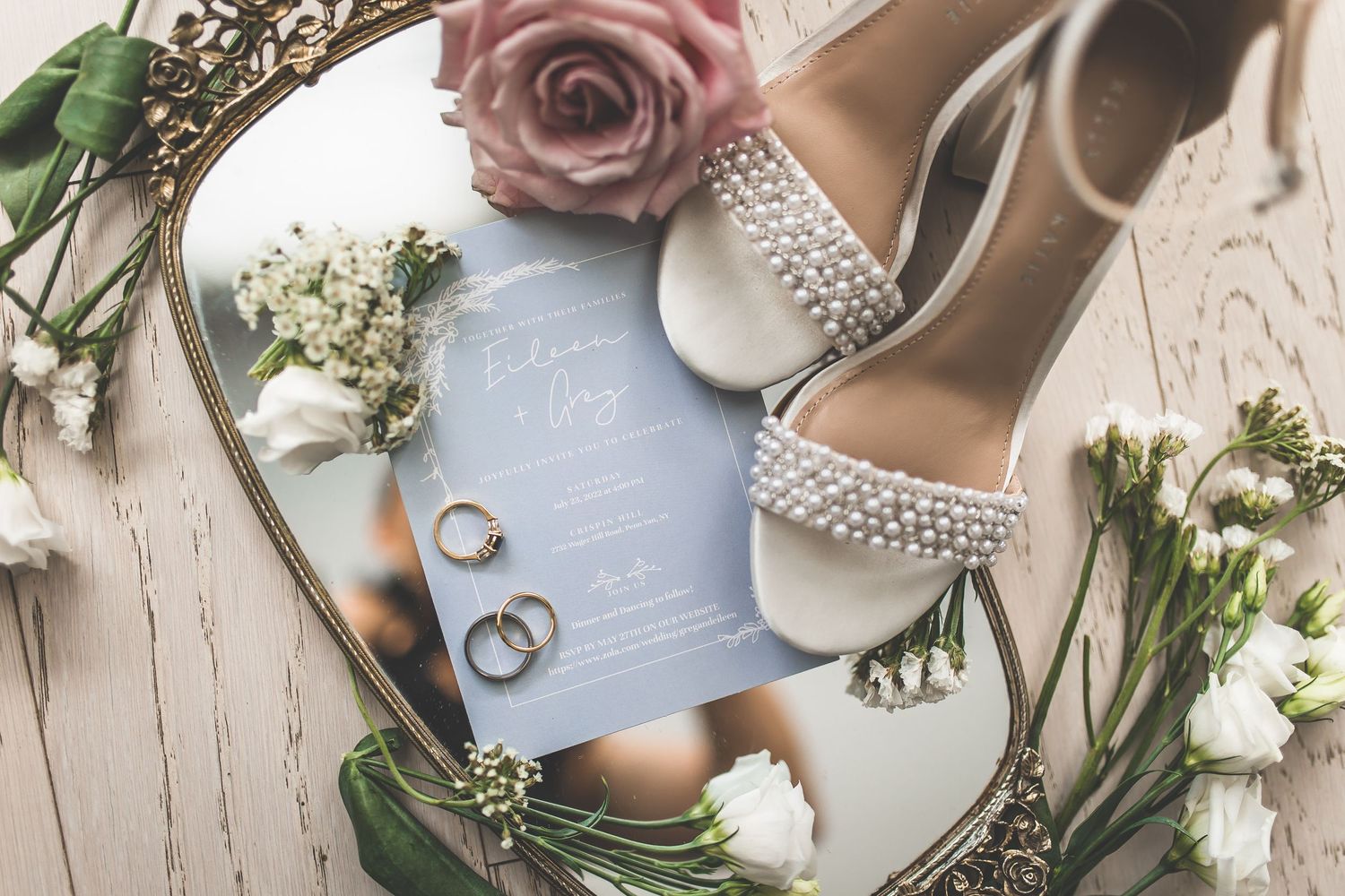 Wedding detail flatlay with dusty rose flowers, rings, and elegant bridal shoes on light background.