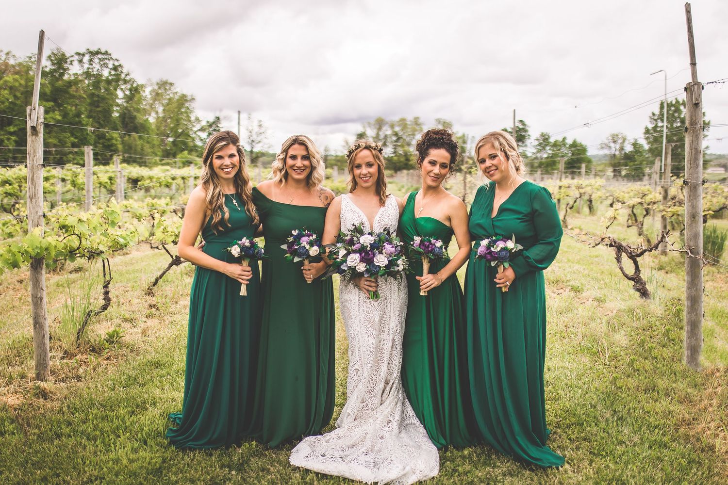 Bridal party in emerald dresses poses among vineyard rows on cloudy day.