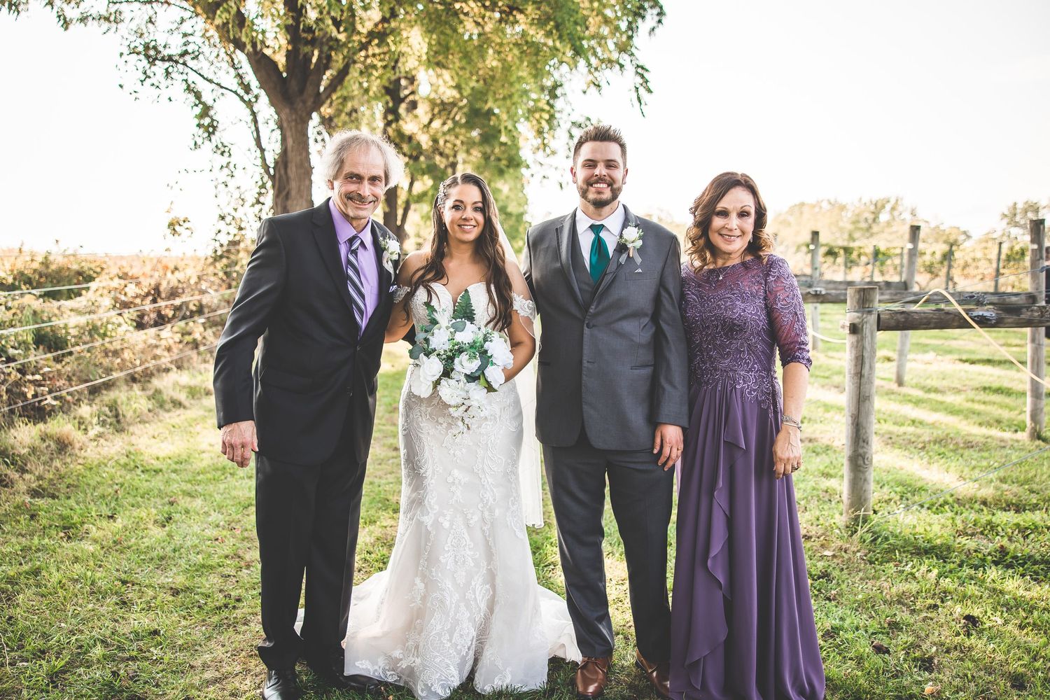 Wedding group photo with attendees in formal attire standing beneath a large tree.