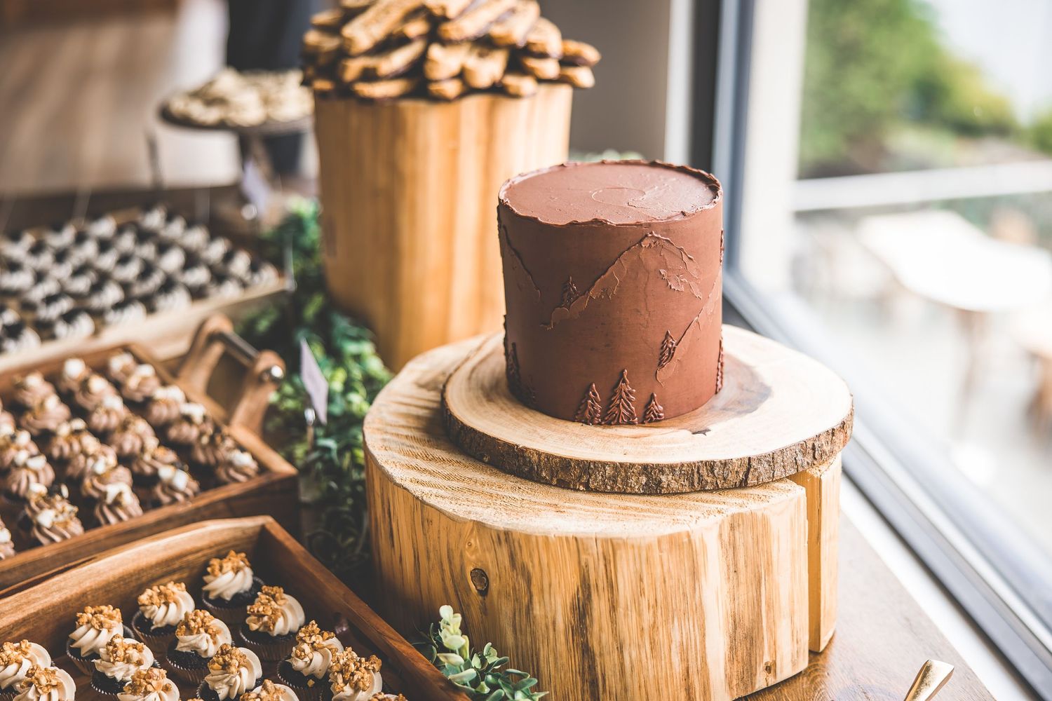 Chocolate wedding cake on wood slice with dessert display near window.