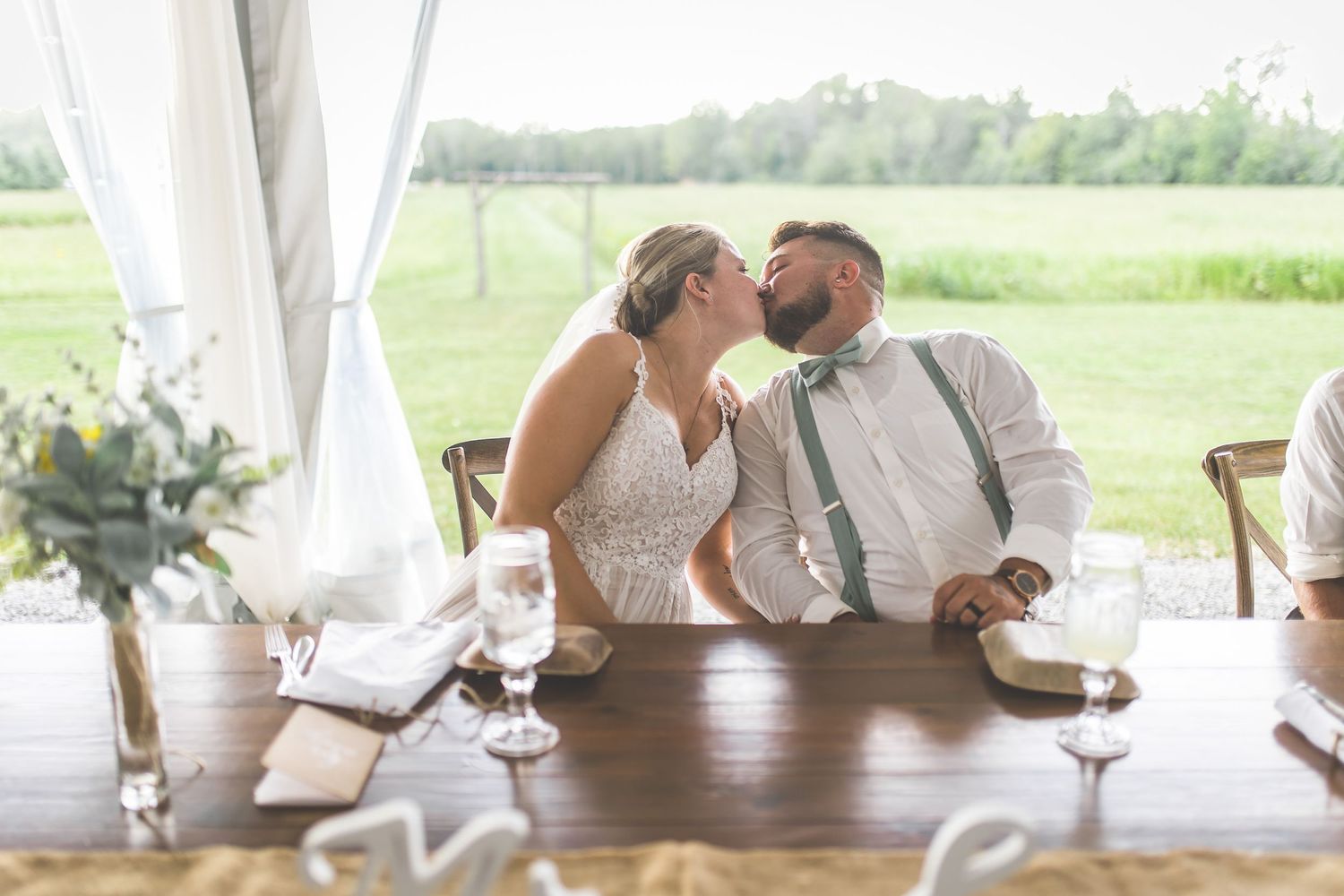 A couple shares an intimate moment at their reception table with rustic outdoor scenery visible.