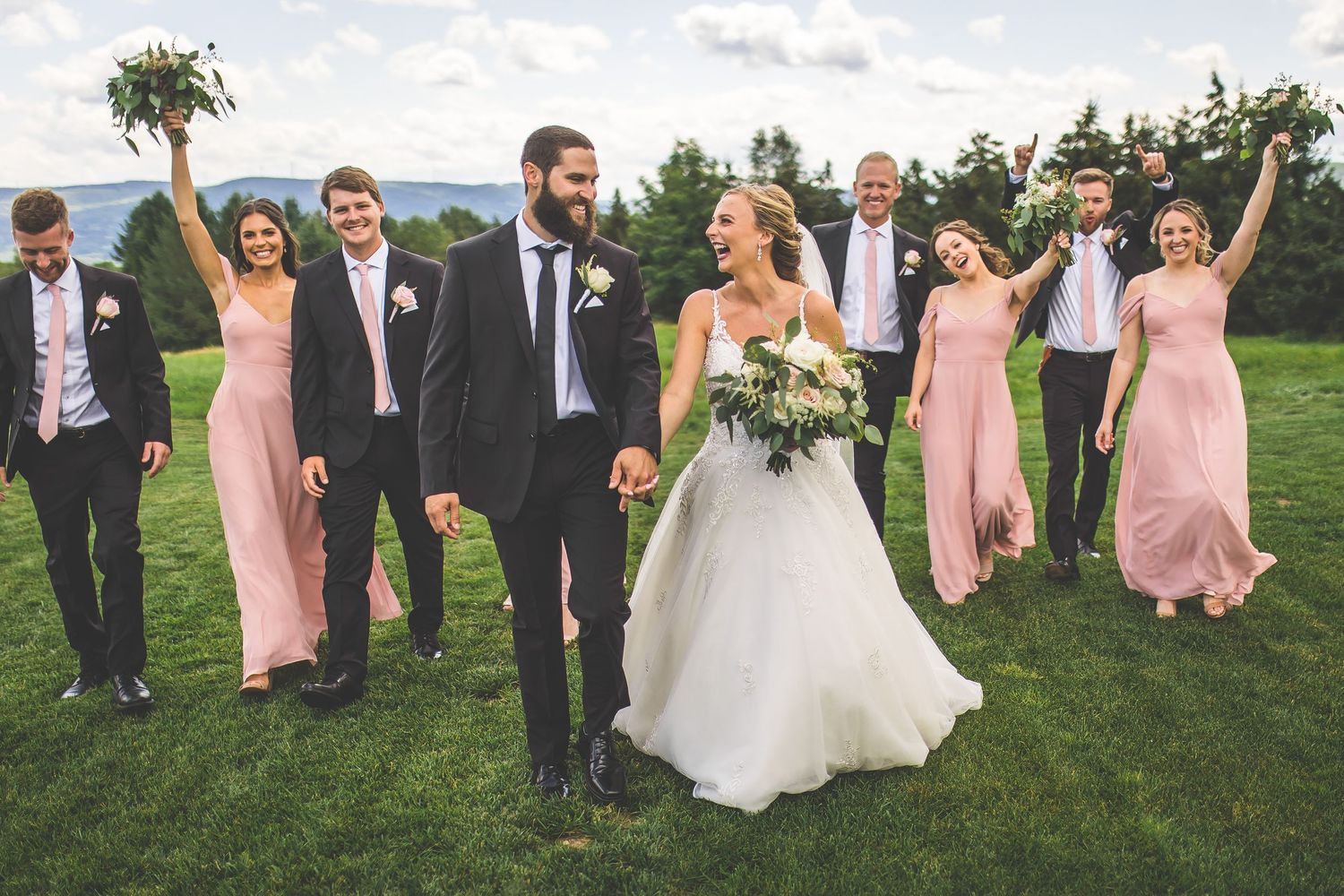 Wedding party celebrates joyfully on grassy field with mountains visible in distance.
