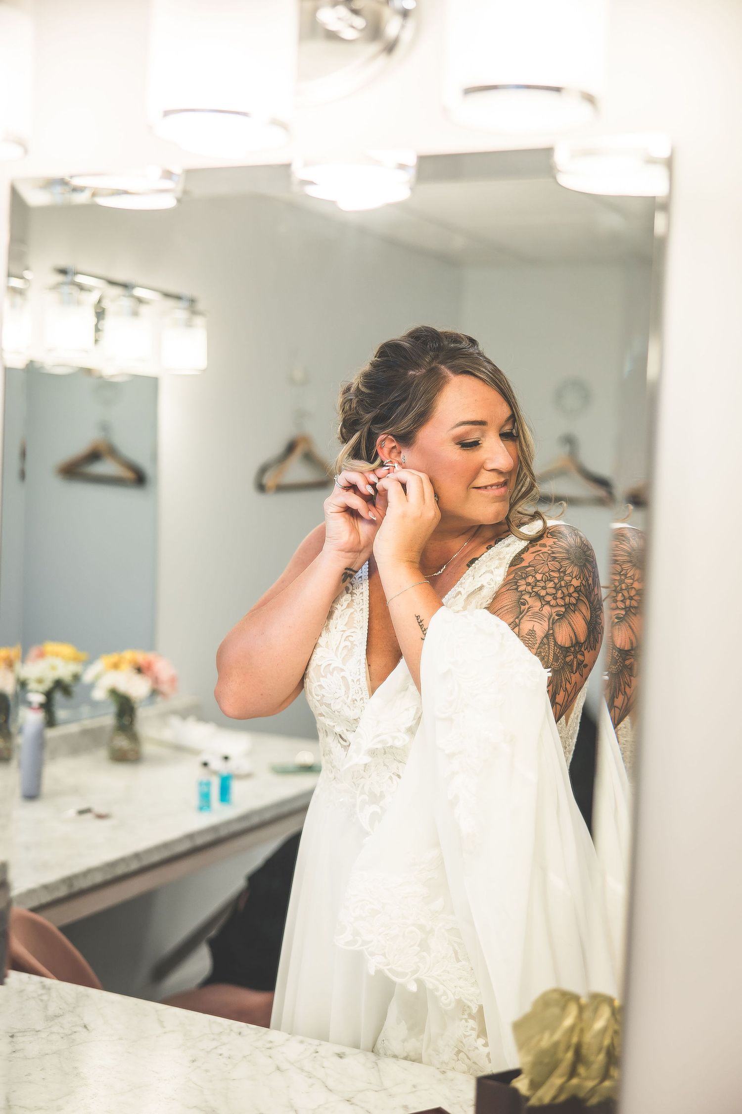 Bride in delicate lace gown prepares in a brightly lit dressing room with mirrors.