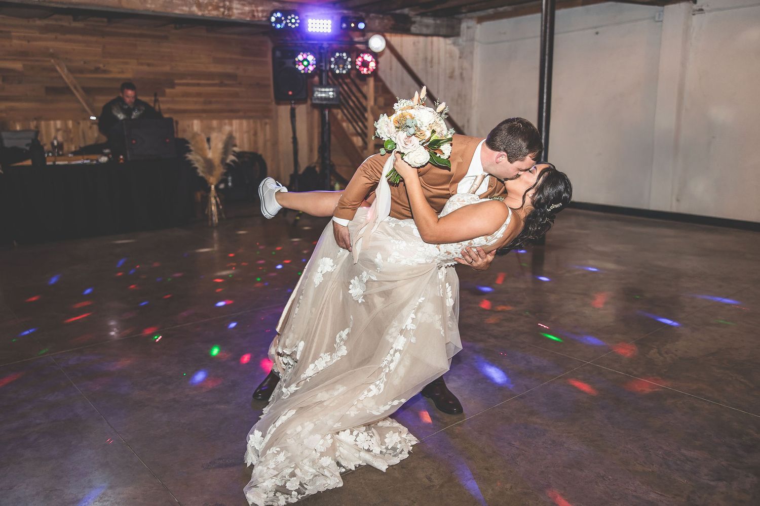 Newlyweds dance under colorful disco lights in a rustic barn venue with wooden beams.