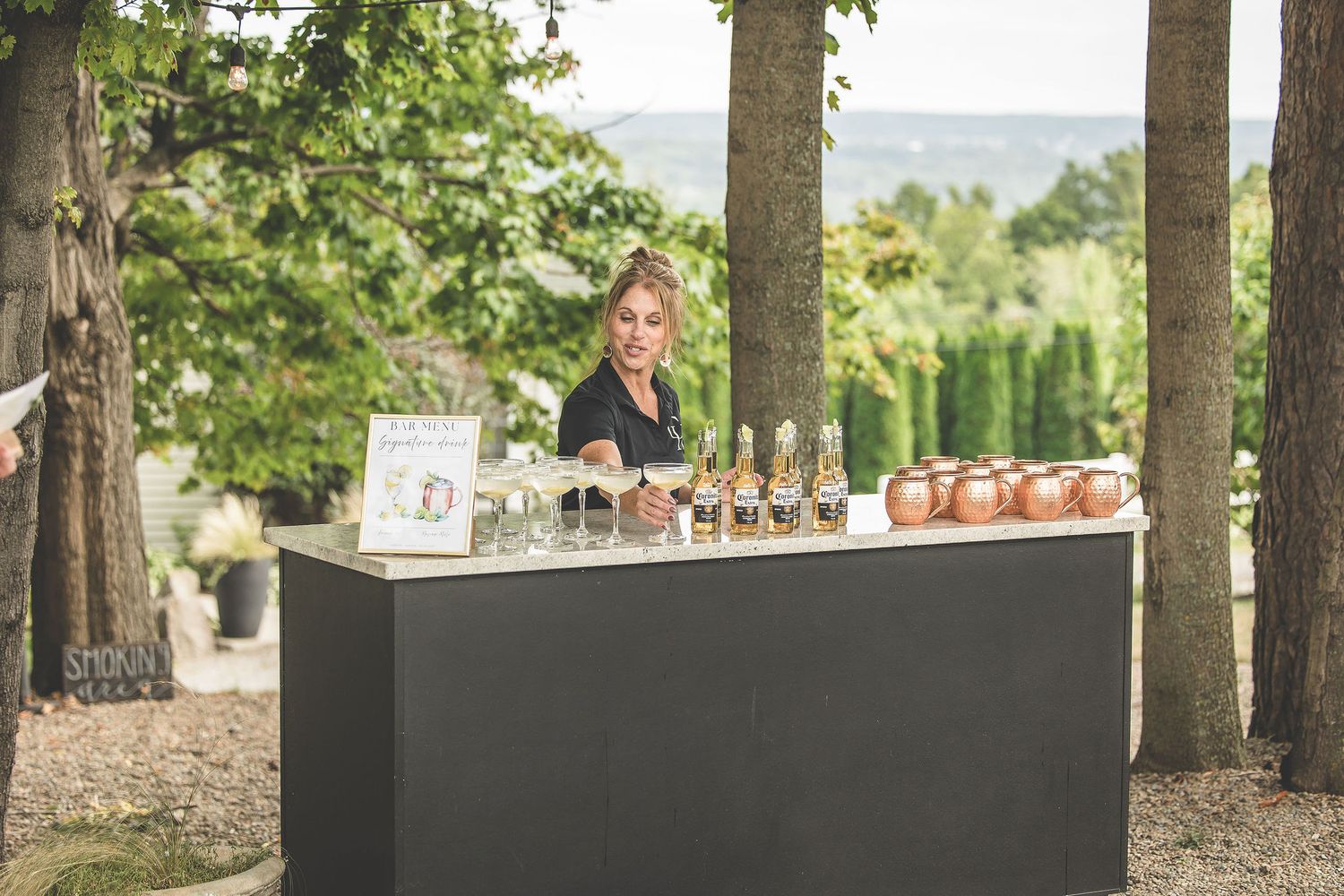 Elegant outdoor bar setup under tall trees with refreshments and glassware displayed.
